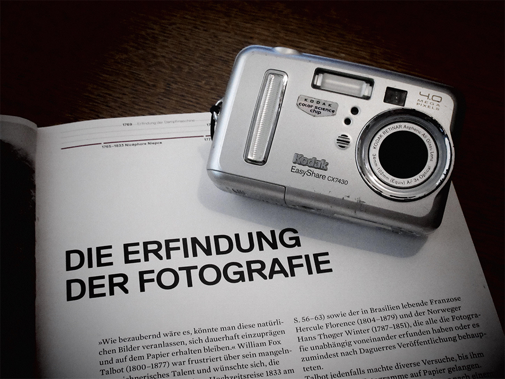 A Kodak digital camera rests on an open German magazine. The visible page headline reads “DIE ERFINDUNG DER FOTOGRAFIE,” which translates to “The Invention of Photography.”
