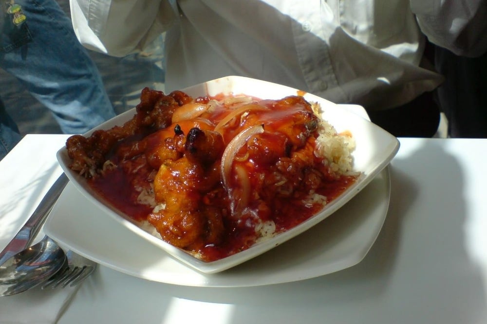 A plate of rice topped with fried chicken pieces, onions, and bright red sweet and sour sauce, placed on a white table next to a fork and spoon.