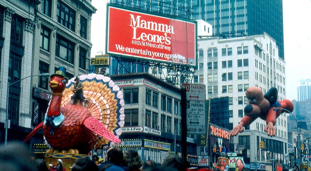 A parade features a large turkey float and a dog balloon, surrounded by crowds in a city. In the background, a red “Mamma Leone’s” restaurant billboard is visible among tall buildings.
