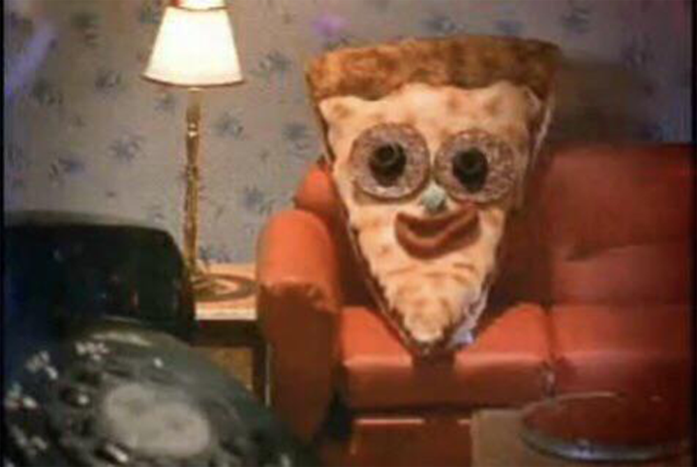 A slice of pizza with human-like features—donut eyes, a red smile, and a nose—sits on a red couch in a living room with a lamp and patterned wallpaper.