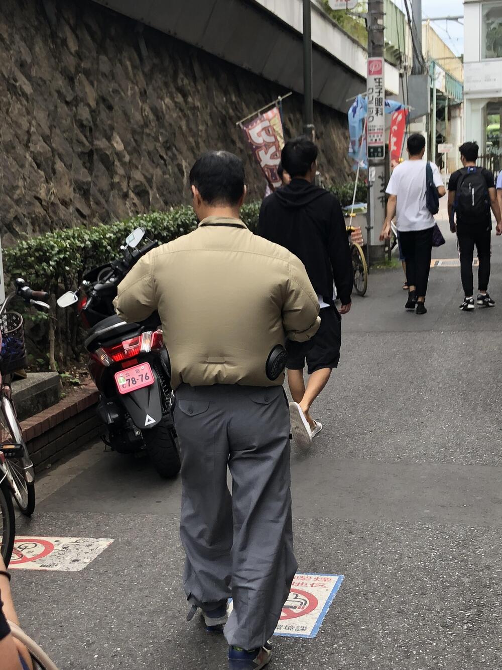 A man wearing a tan jacket with built-in fans walks down a city street; other people walk ahead, and motorcycles are parked along the sidewalk.