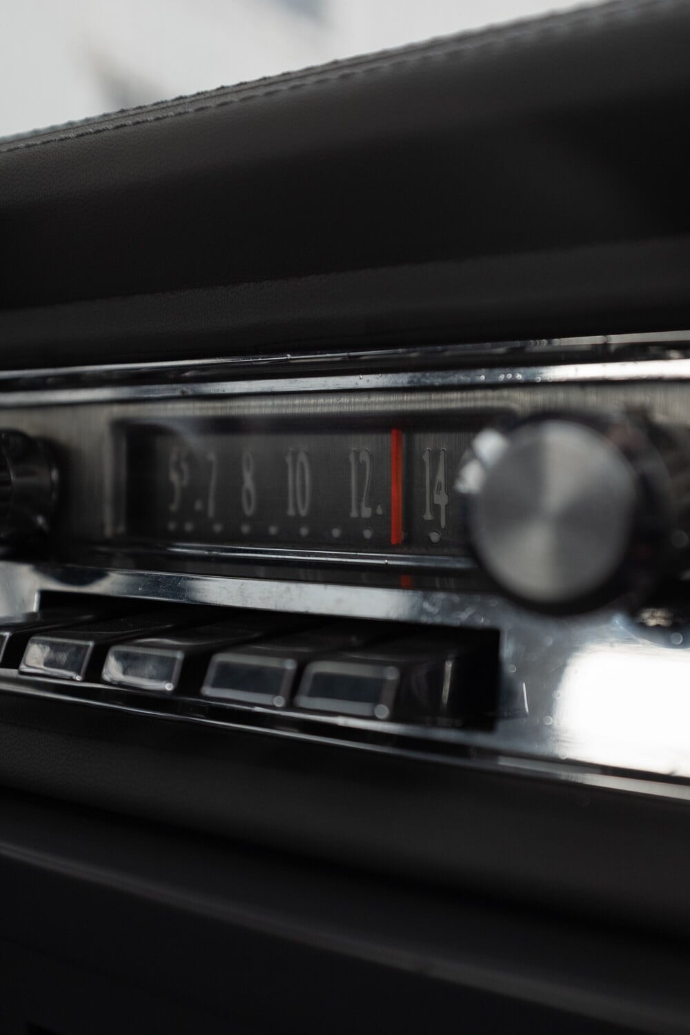 Close-up of a vintage car radio showing frequency numbers and silver control knobs, with a red indicator pointing around 12, and several buttons below the display.