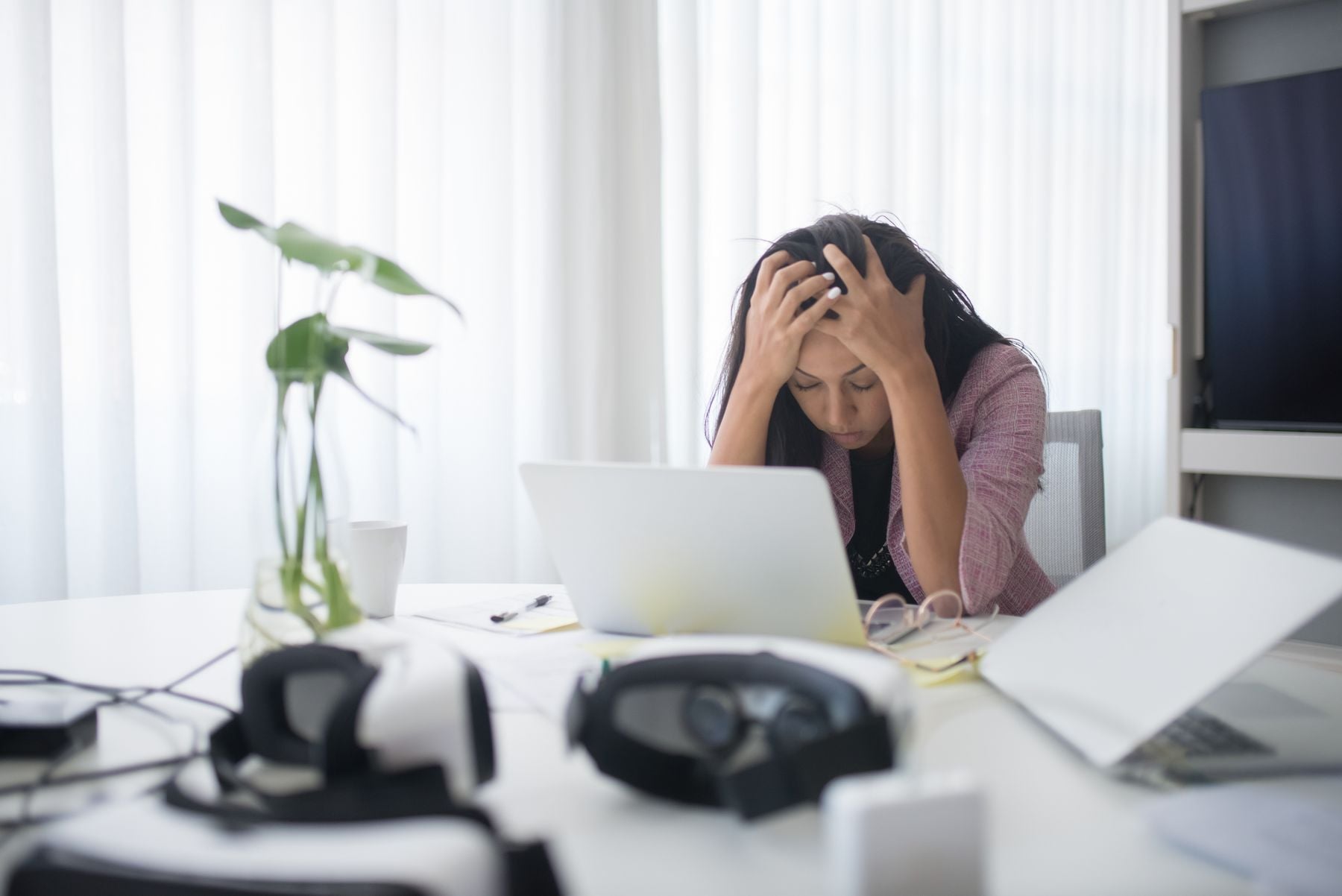 A person sits at a desk, holding their head in their hands with frustration. A laptop, papers, and VR headsets are on the desk. Bright light filters through sheer curtains behind them.