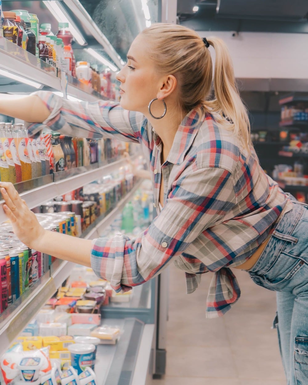 A woman in a plaid shirt and jeans reaches for a drink on a refrigerated shelf in a grocery store, with various drinks and food items visible around her.