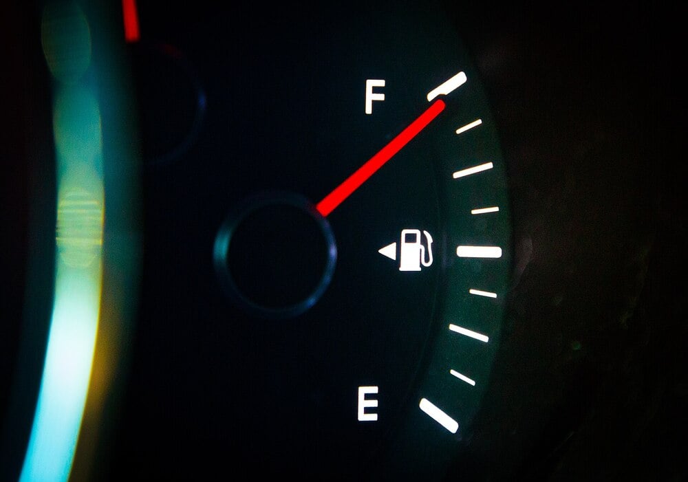 A close-up of a car’s fuel gauge showing a full tank. The needle points to “F,” and a fuel pump icon appears near the center of the dial. The gauge is illuminated in dark surroundings.