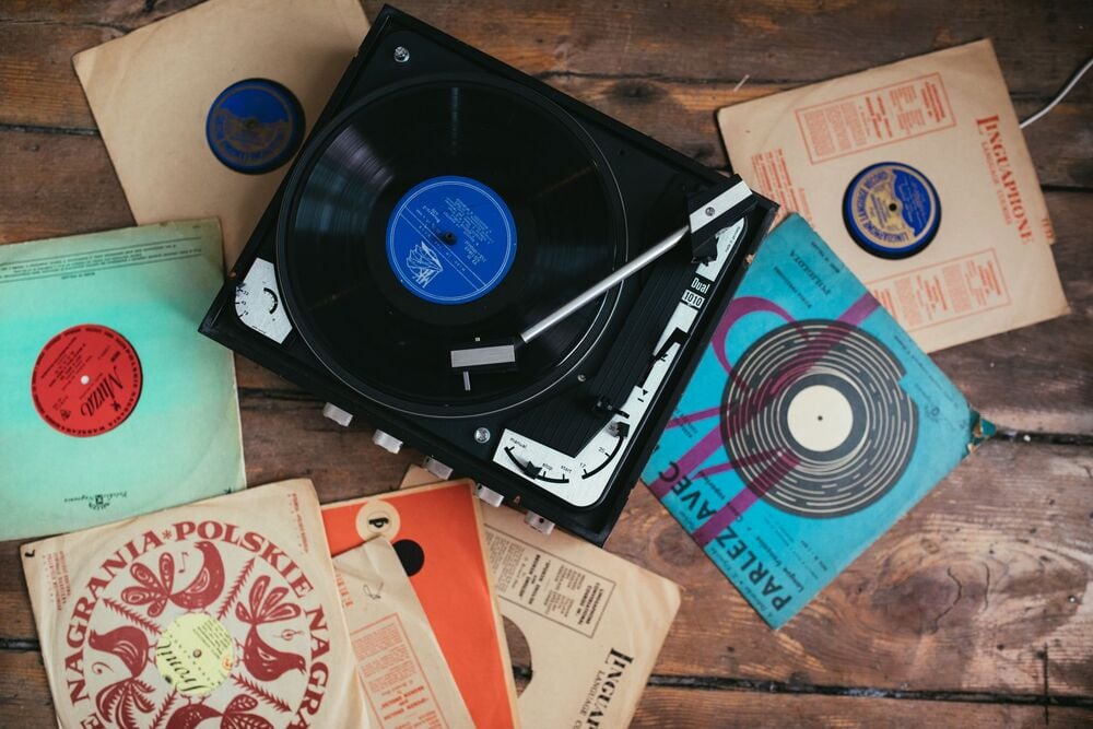 A black record player with a vinyl record on it sits on a wooden floor, surrounded by vintage record sleeves and more vinyl records in various colors and designs.