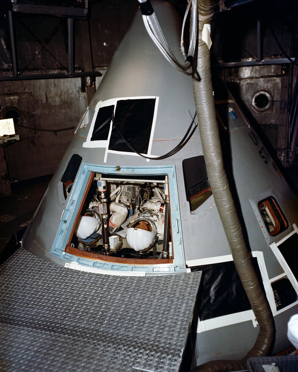 Two astronauts in white spacesuits are seated inside a gray Apollo command module with its hatch open, surrounded by cables and metal platforms inside a spacecraft facility.