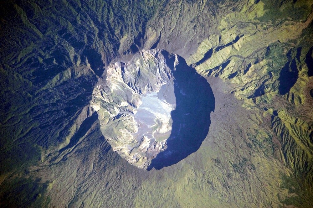 An aerial view of a large volcanic crater surrounded by green forested slopes and ridges, with the crater’s rocky interior clearly visible.