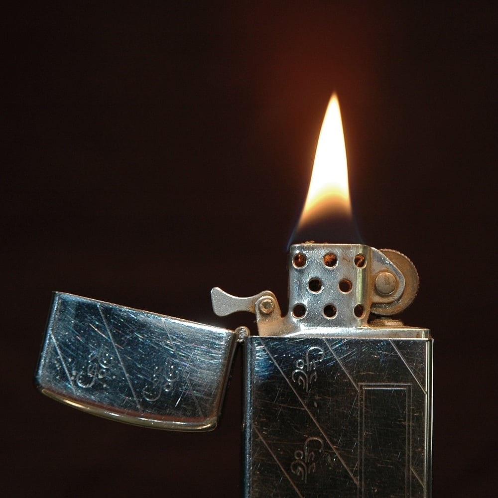 A close-up of a metal lighter with its lid open, producing a steady flame against a dark background.