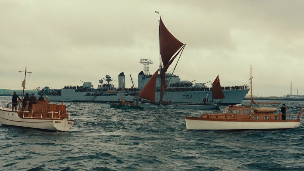 Several small wooden boats and a sailboat gather near a large navy warship at sea under a cloudy sky, with people visible on all vessels.