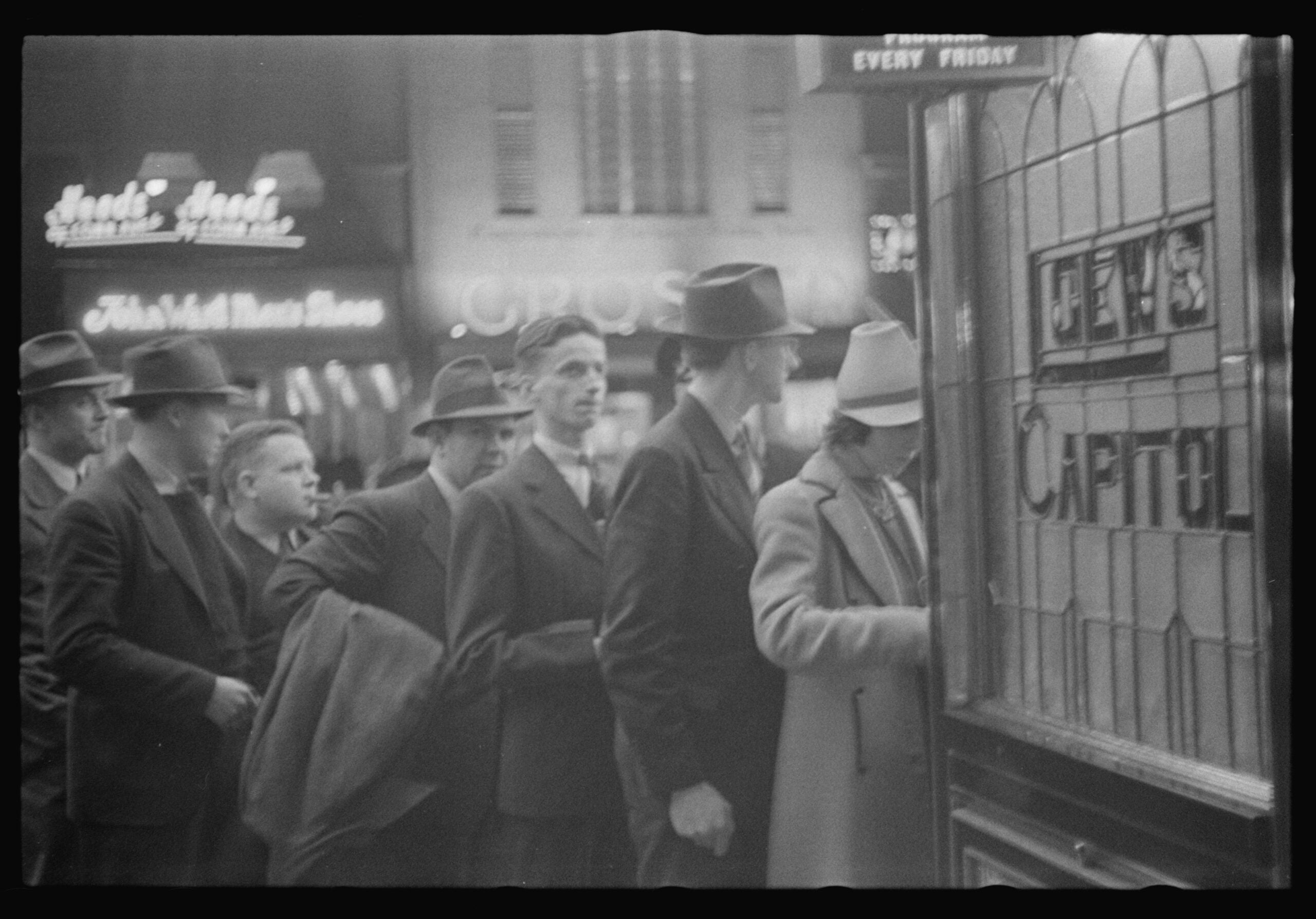 Black-and-white photo of men in suits and hats standing in line at a ticket booth outside a theater at night. Neon signs and theater lights are visible in the background.