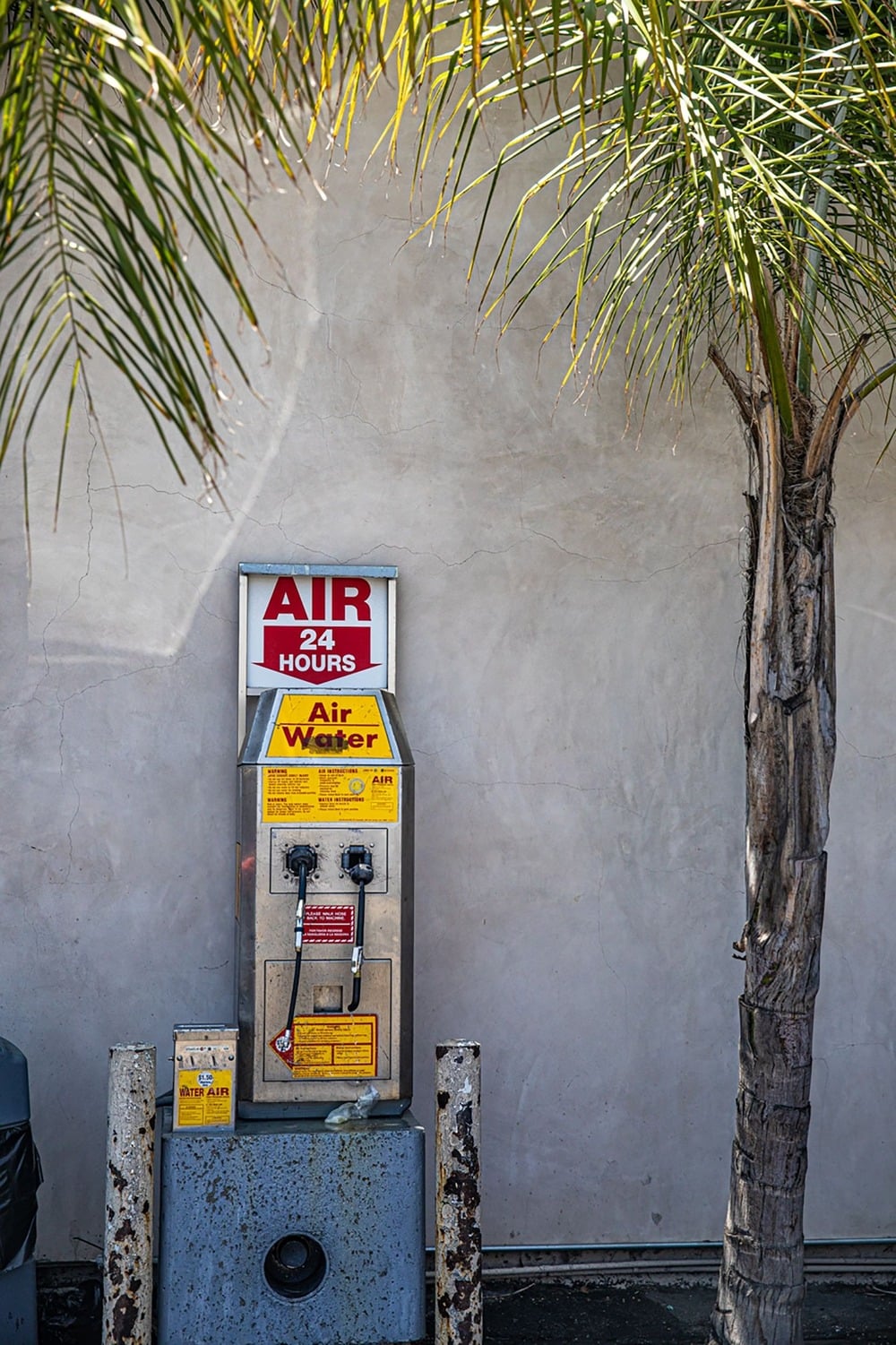 A public air and water machine stands by a weathered wall, partially shaded by palm tree fronds, with signs for “Air 24 Hours” and instructions above the coin-operated valves.