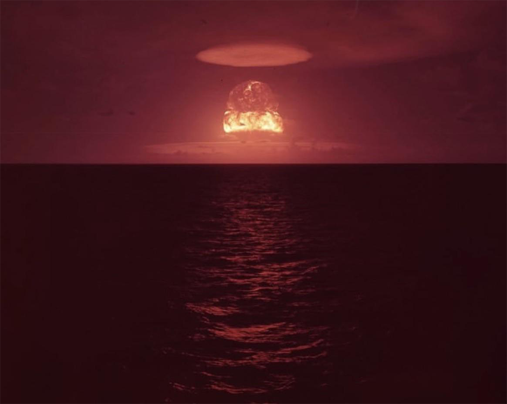 A large, fiery mushroom cloud rises over the ocean at dusk, casting a red-orange glow on the water’s surface beneath a dark, cloudy sky.
