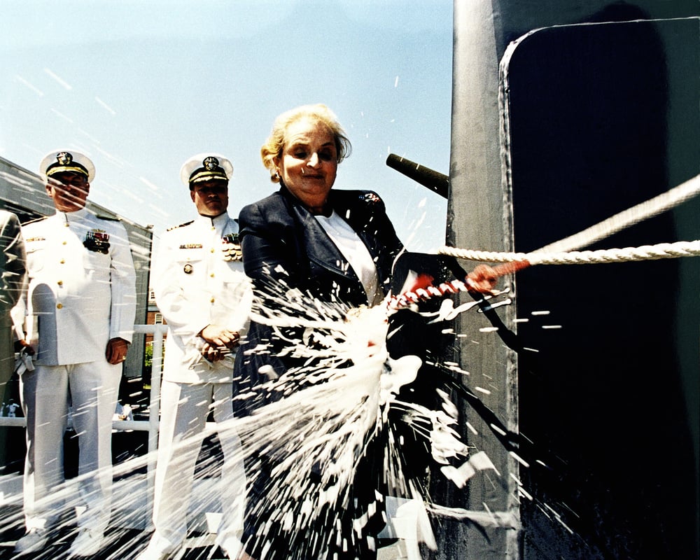 A woman in a suit breaks a champagne bottle against a naval vessel as two uniformed officers watch, during a ship christening ceremony. Liquid splashes outward from the impact.