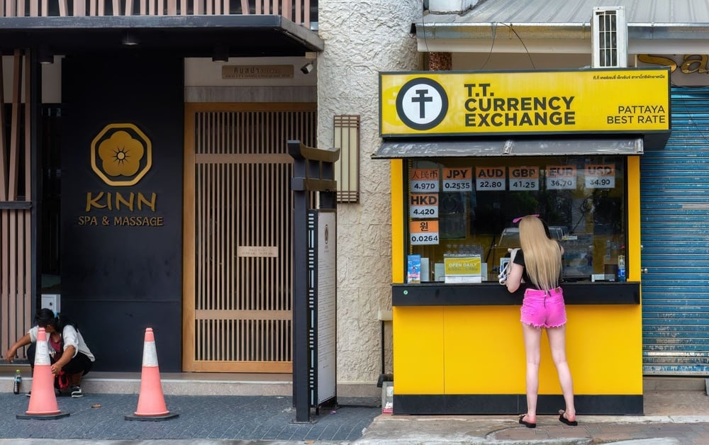 A woman in pink shorts stands at a yellow T.T. Currency Exchange booth next to the entrance of Kinn Spa & Massage. Another person sits on the sidewalk near two traffic cones.