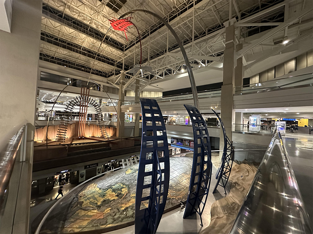 Large modern airport atrium with tall metal sculptures, an illuminated world map on the floor, and stores in the background. High ceilings and bright lights create an open, spacious atmosphere.
