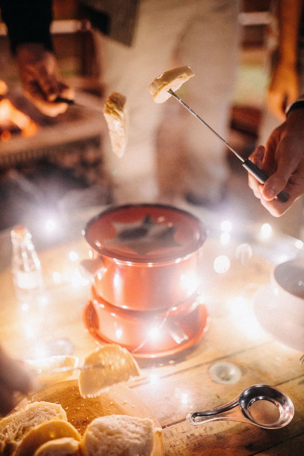 People dip bread into a red fondue pot on a wooden table, surrounded by string lights and utensils, creating a cozy and festive atmosphere.