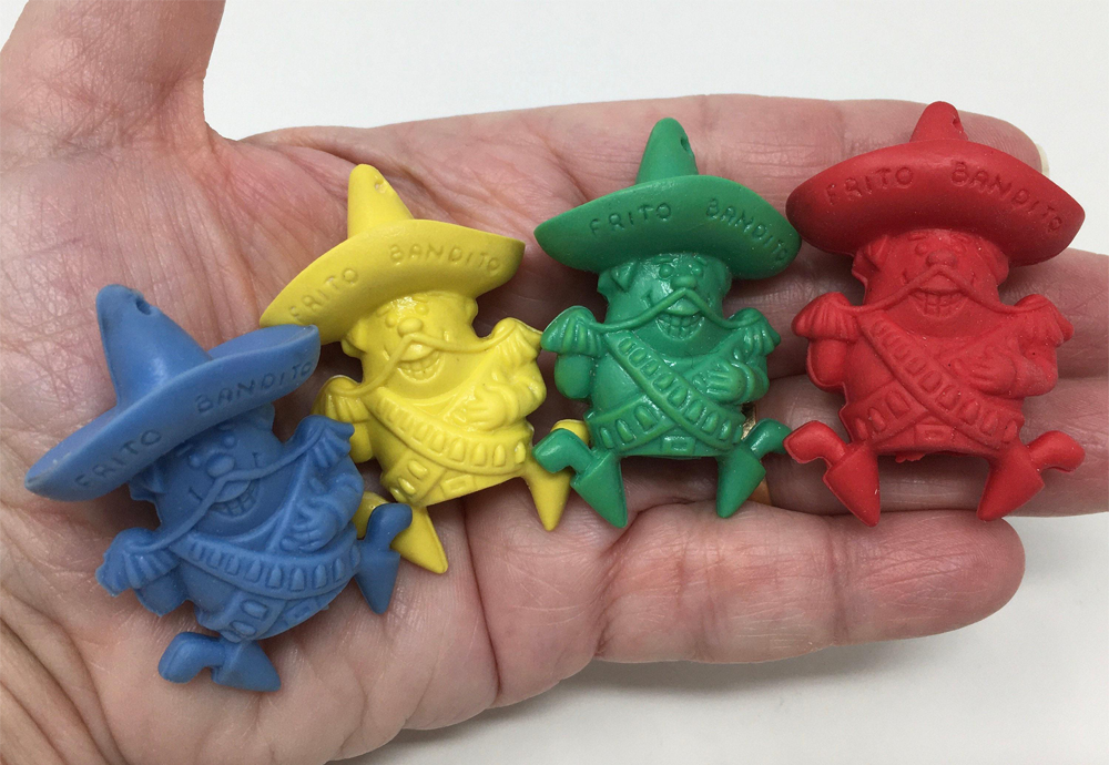 A hand holds four plastic Frito Bandito pencil toppers in blue, yellow, green, and red, each wearing a sombrero and sash, and smiling with a mustache.
