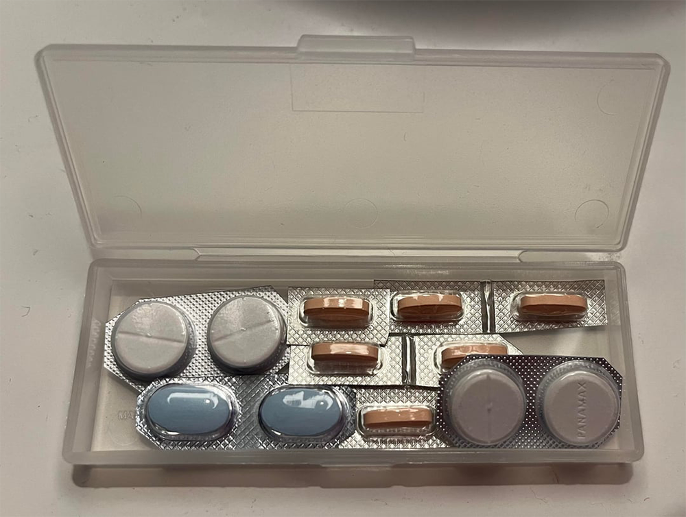 A plastic pill case holds several blister packs with oval pink, round white, and oval blue pills arranged neatly inside.