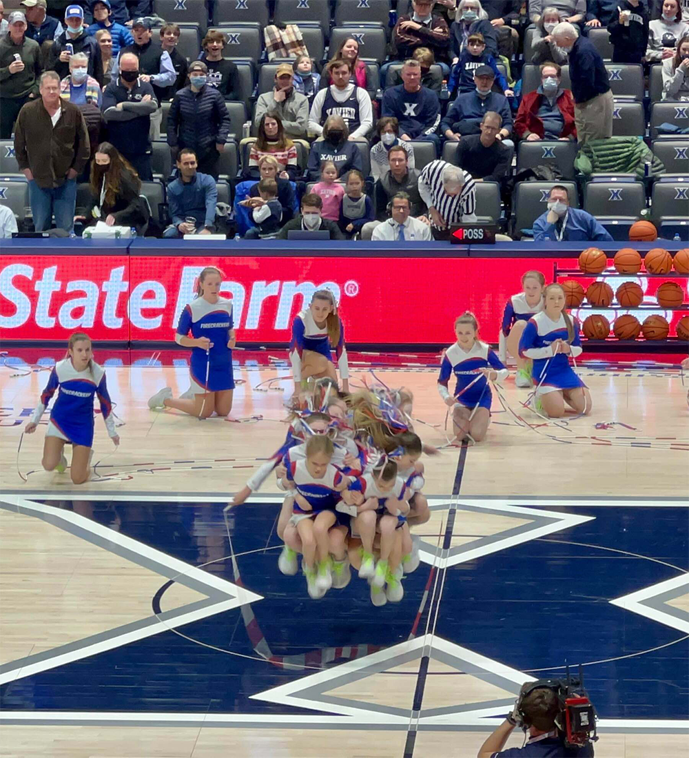 A cheerleading team in blue, white, and red uniforms performs a synchronized jump rope routine on a basketball court, with a crowd watching from the stands.