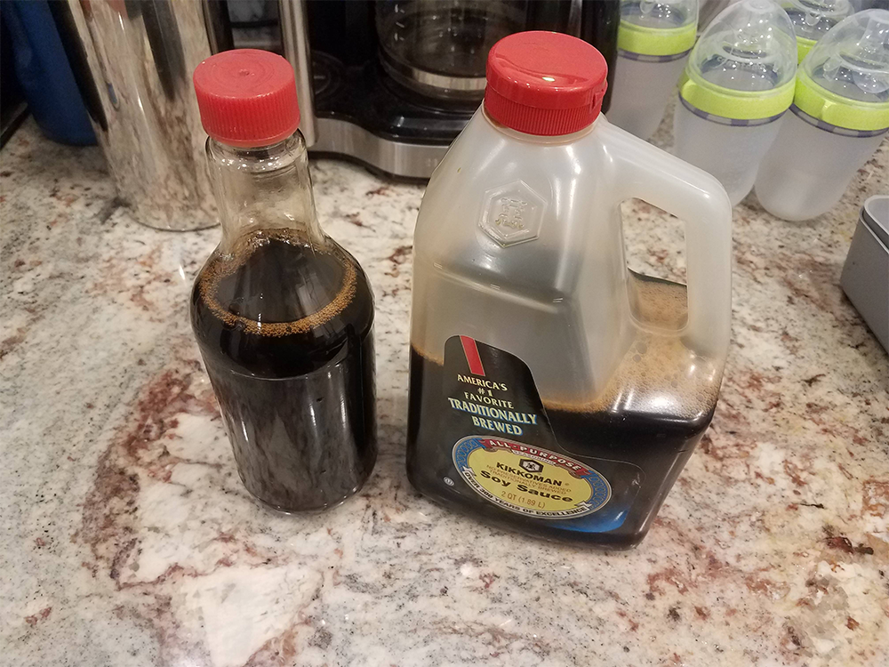 A glass bottle and a large plastic jug, both filled with dark soy sauce, sit on a kitchen counter near a coffee maker and several baby bottles.
