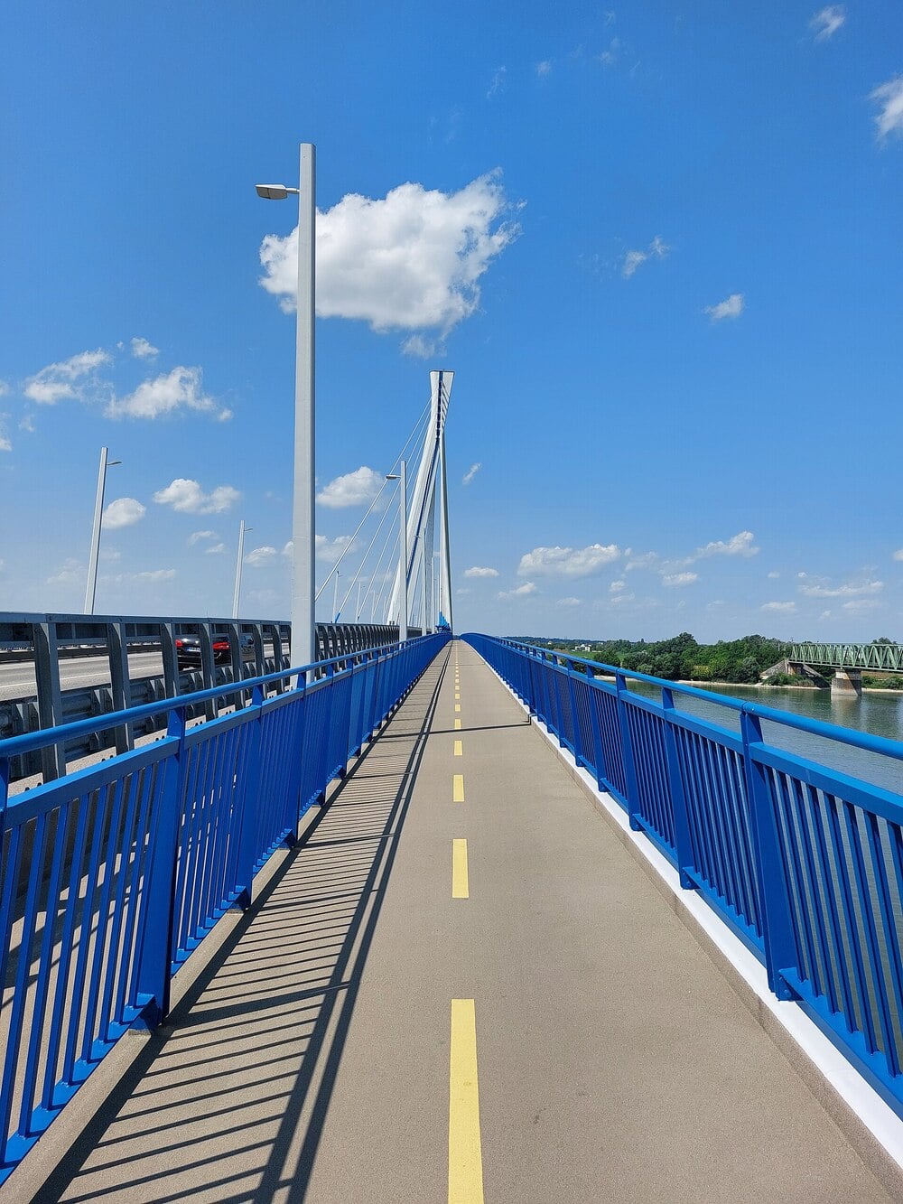 A straight pedestrian and bike path with bright blue railings crosses a modern bridge over water under a sunny, clear blue sky with a few clouds.