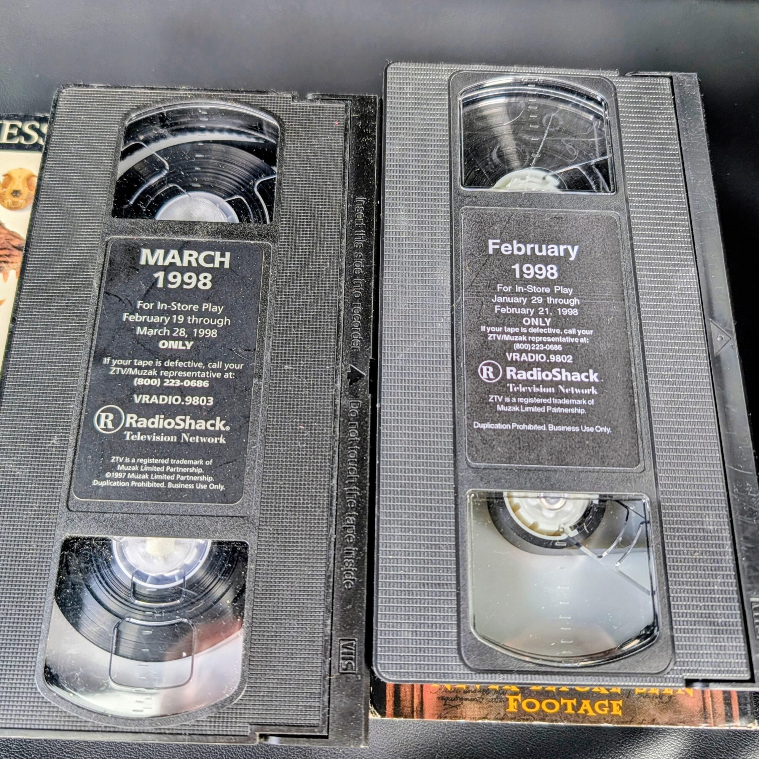 Two VHS tapes labeled “March 1998” and “February 1998” from RadioShack sit side by side, partially overlapping, on top of other video cassette covers.