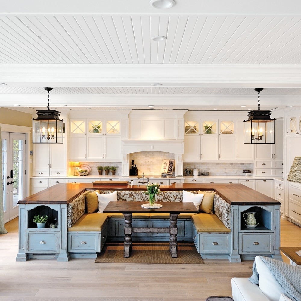 A bright kitchen with white cabinets, a large island featuring a built-in bench and table, wooden countertops, and two black pendant lights hanging above the island.