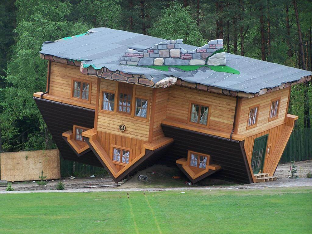A wooden house built upside down, with its roof on the ground and foundation in the air, surrounded by trees and grass. The windows, doors, and roof appear inverted.