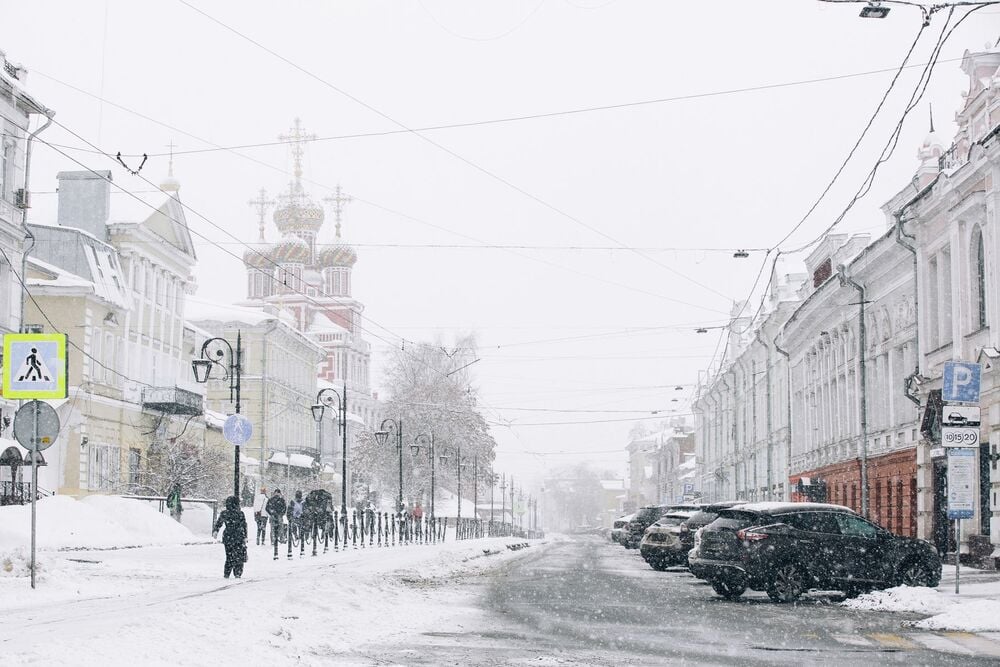 Snow-covered city street with parked cars, people walking on the sidewalk, ornate church with golden domes in the background, and old buildings lining both sides under falling snow.