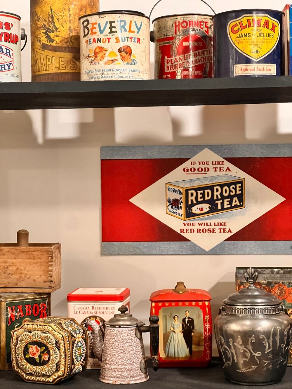A display of vintage tins and teapots on shelves, featuring colorful labels for peanut butter, syrup, tea, and other goods, with a Red Rose Tea sign in the center and a decorative teapot and photo in front.