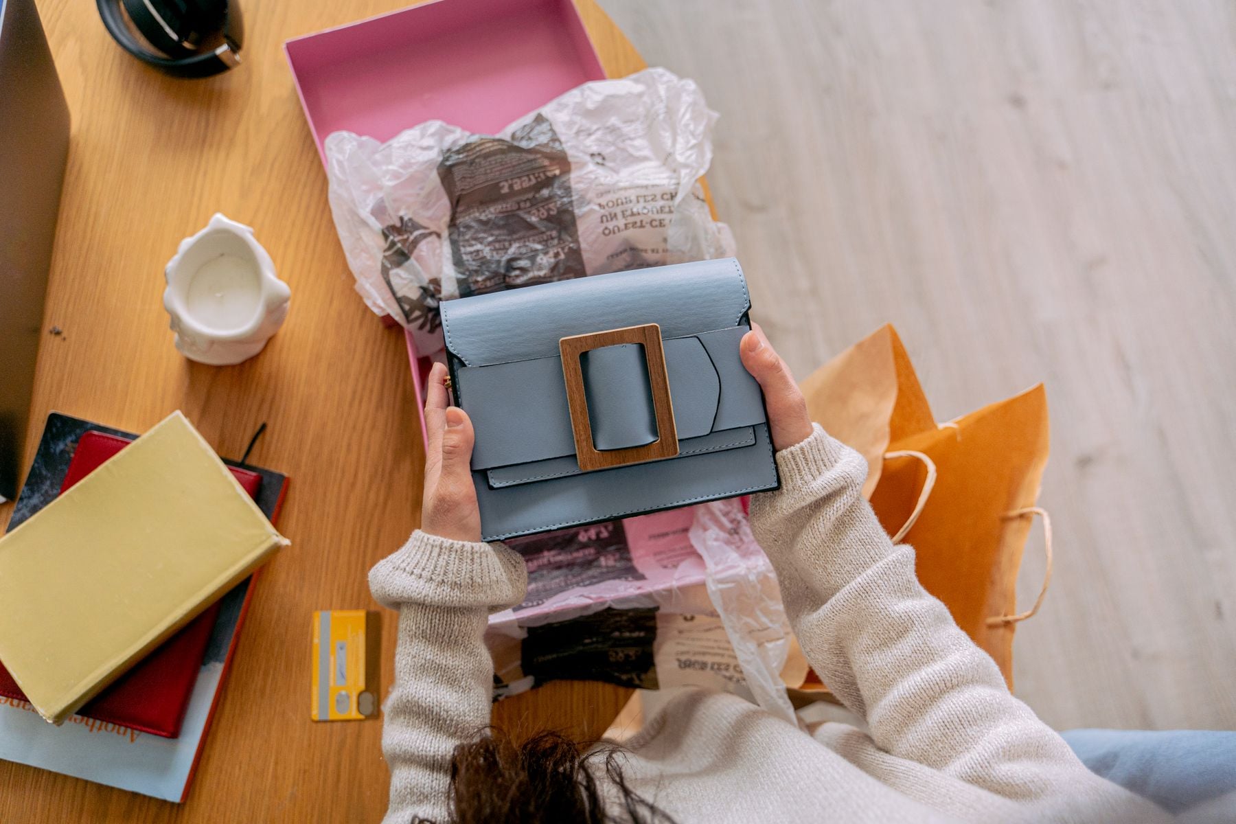 A person holds a light blue handbag with a large square buckle over a wooden table, surrounded by books, a candle, a yellow credit card, and shopping bags.