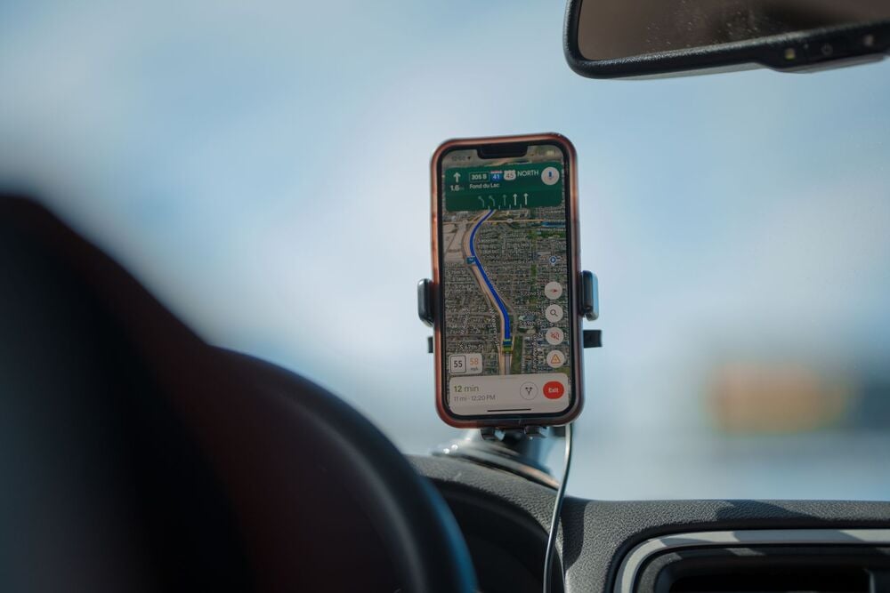 A smartphone mounted on a car dashboard displays a GPS navigation map with a planned driving route on the screen. The phone is connected with a cable, and part of the windshield is visible.