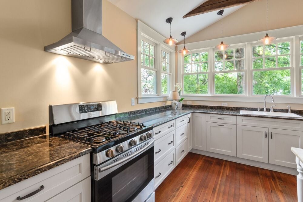 Bright kitchen with white cabinets, stainless steel stove and hood, marble countertops, wood floors, pendant lights, and large windows showing green trees outside.