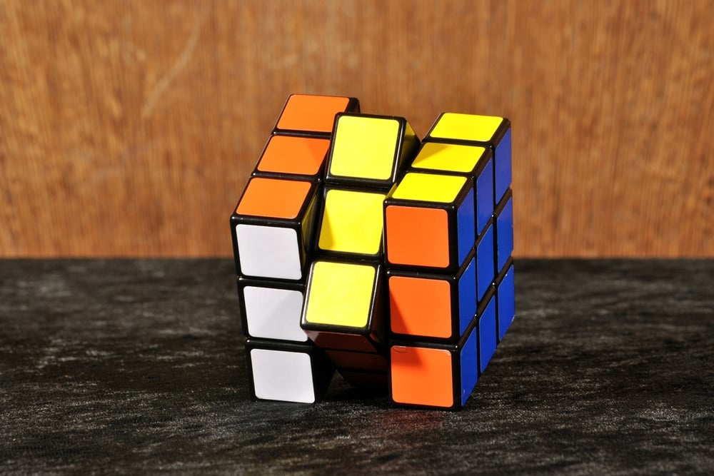 A partially solved Rubik’s Cube with orange, yellow, blue, and white faces is sitting on a dark textured surface, against a wooden background. The cube is twisted, with some squares misaligned.