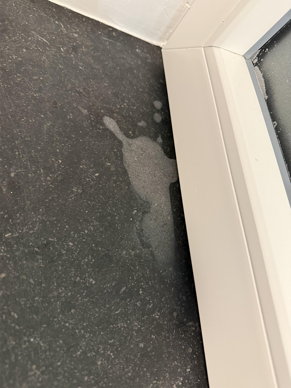 A wet patch or stain on a dark floor near the corner of a white window frame.