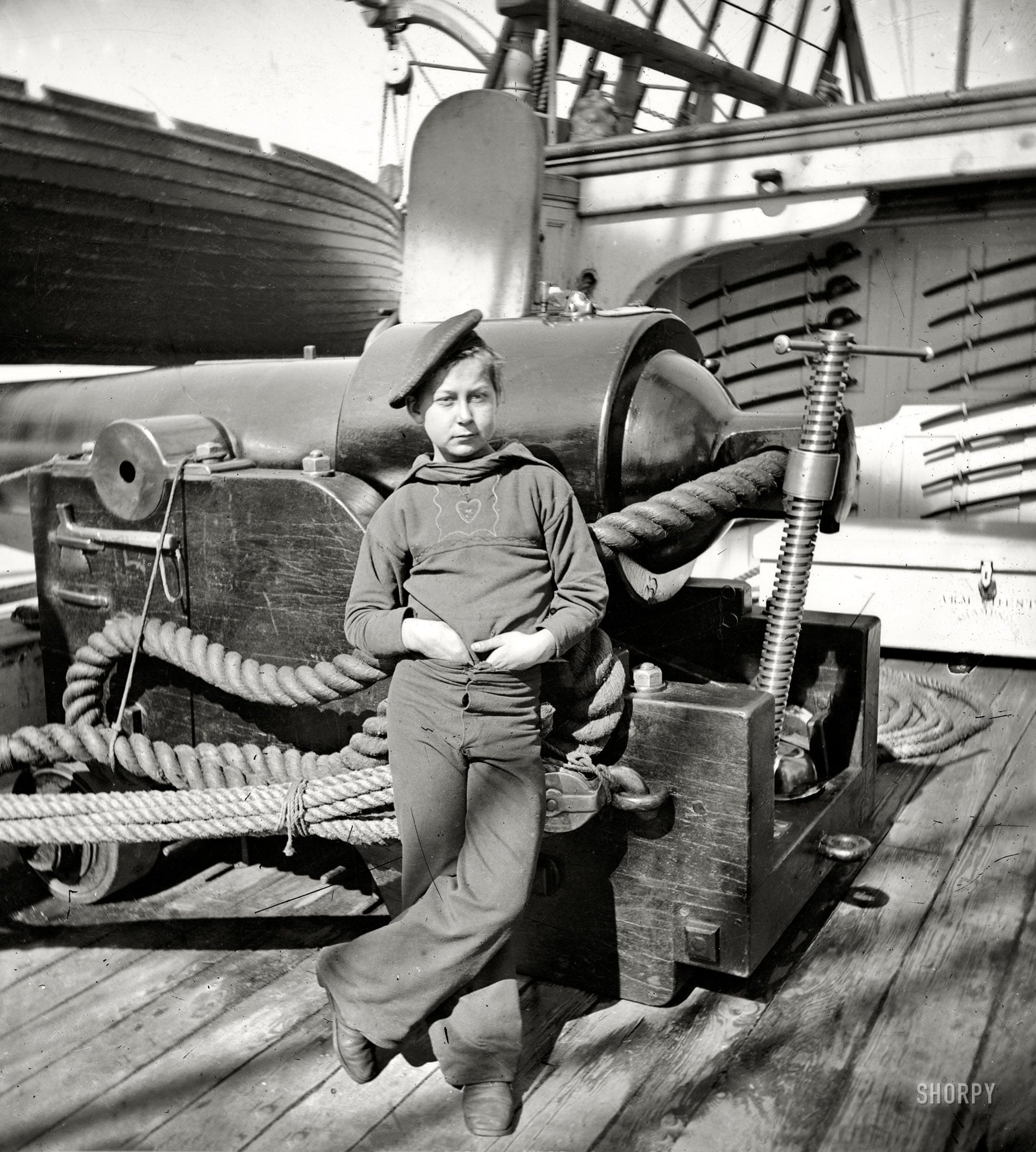 A young sailor in naval uniform leans against a large cannon on a ship deck, surrounded by coiled ropes and nautical equipment, with lifeboats and rigging visible in the background.
