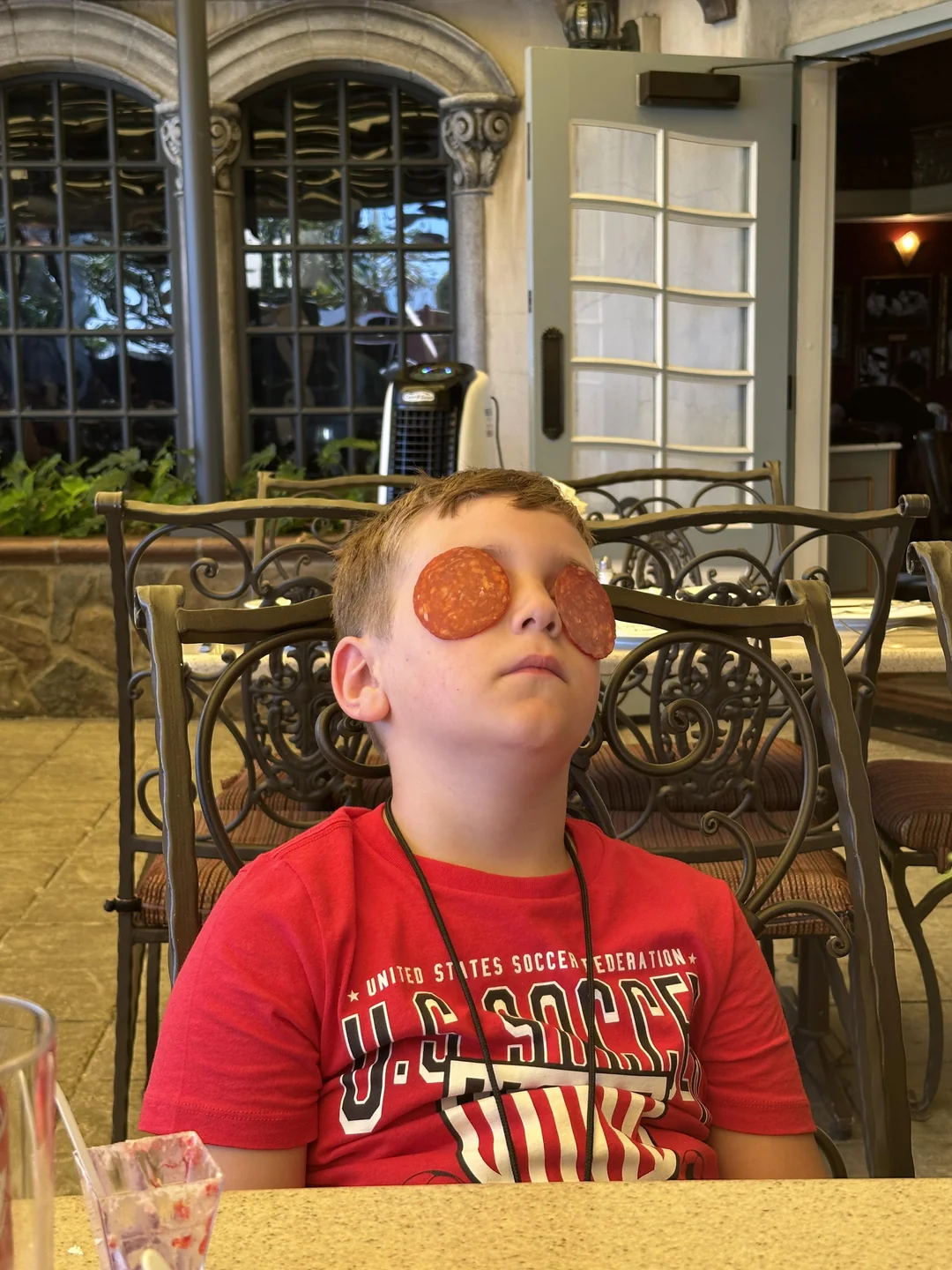 A boy in a red shirt sits with his eyes closed, and slices of pepperoni covering his eyelids, at an outdoor restaurant table with ornate chairs and large windows in the background.