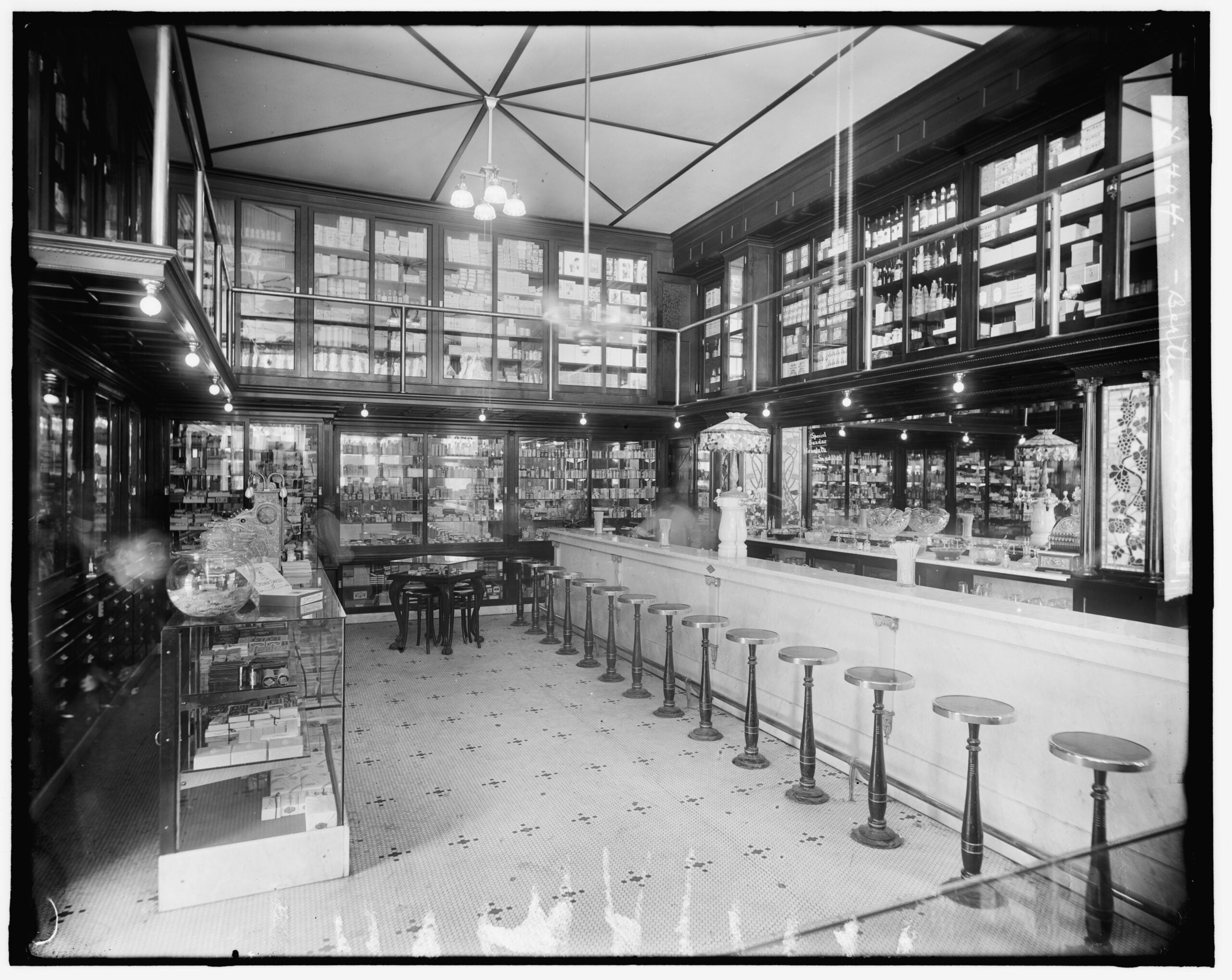Black and white photo of an old-fashioned pharmacy or soda fountain with a marble counter, stools, glass display cases, and shelves filled with bottles and jars.
