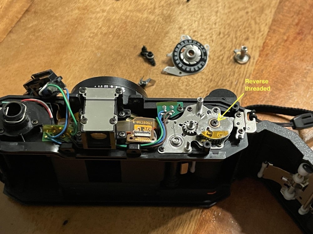 An open film camera showing internal gears and wires on a wooden surface. Several small parts are disassembled nearby. A yellow arrow points to a gear labeled “Reverse threaded” with text overlay.