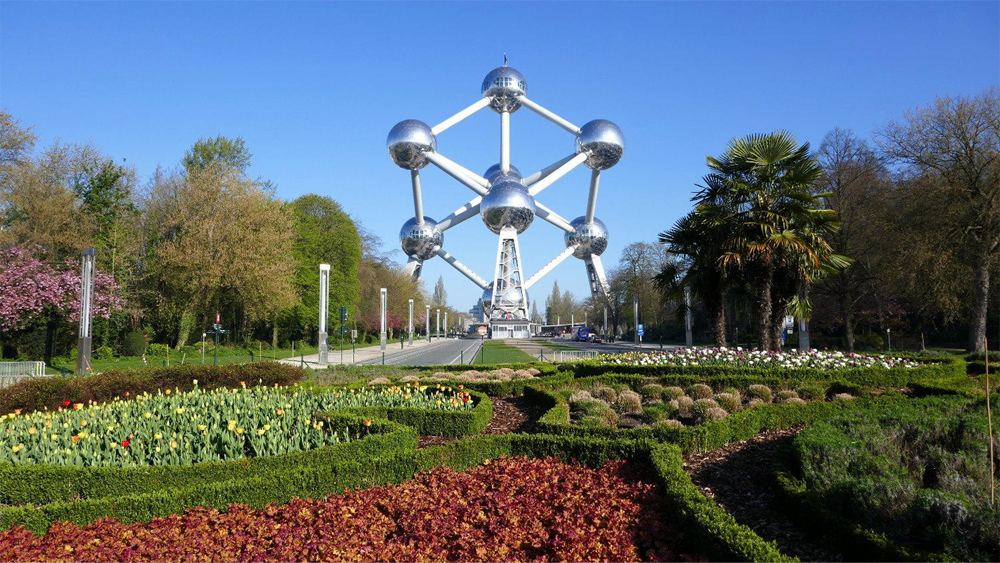 The Atomium in Brussels, Belgium, stands tall behind a landscaped garden with colorful flowers and neatly trimmed bushes under a clear blue sky.