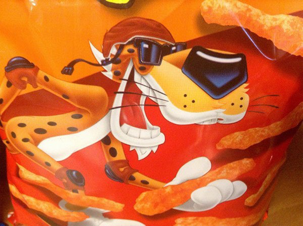 Cartoon cheetah wearing sunglasses and a hat, grinning with an open mouth, surrounded by puffy orange cheese snacks on a bright orange snack bag.