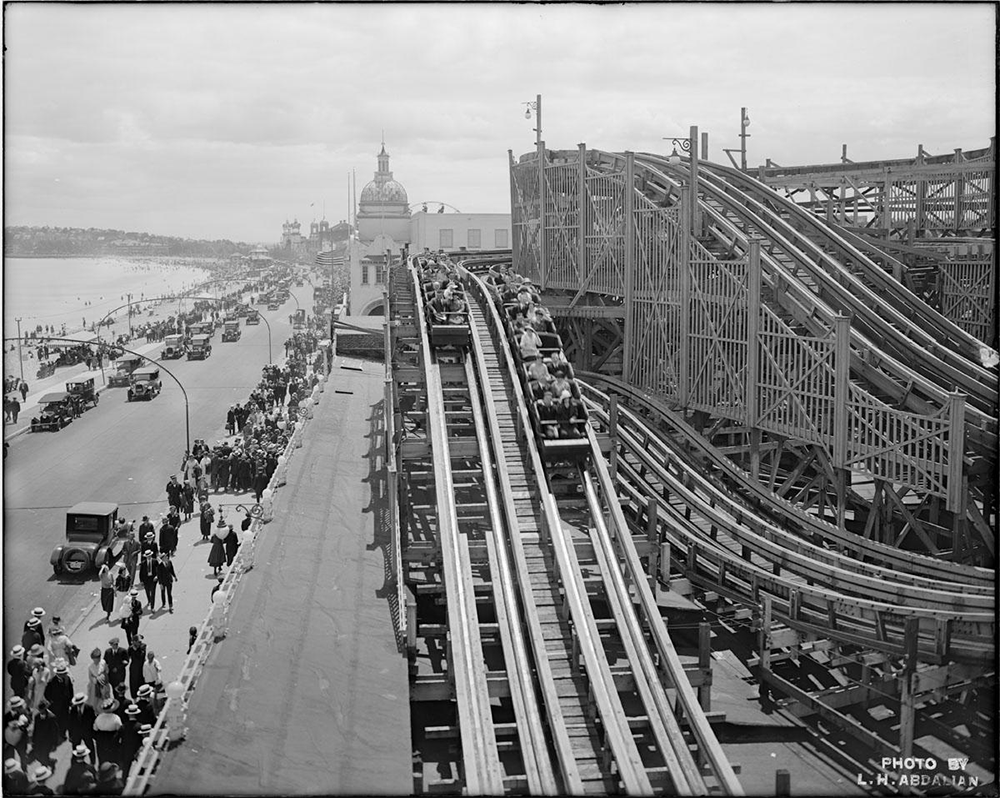 A black-and-white photo shows a crowded wooden roller coaster by a busy seaside road, with people walking and vintage cars driving along the shore. The ocean and large buildings are visible in the background.