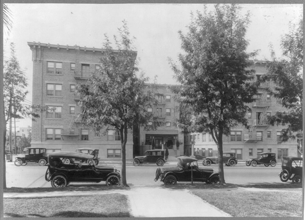 Black-and-white photo of vintage cars parked along a street in front of a four-story brick apartment building with trees lining the sidewalk.