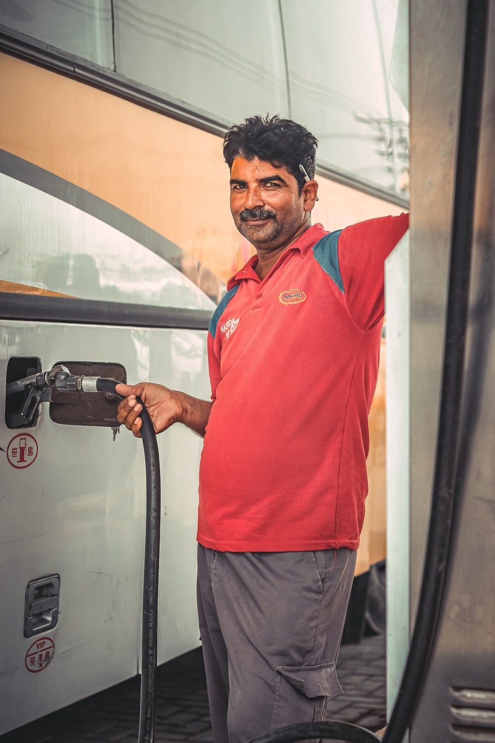 A man in a red shirt smiles while refueling a bus at a gas station. He holds the fuel nozzle with one hand and stands next to the bus, which is white and yellow.