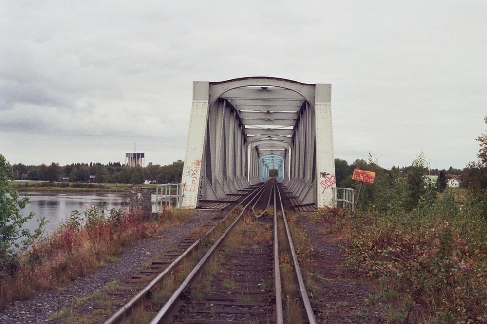 Railroad tracks lead into a metal truss bridge over a river, with some graffiti on the bridge and overgrown greenery on the right side. The sky is overcast.