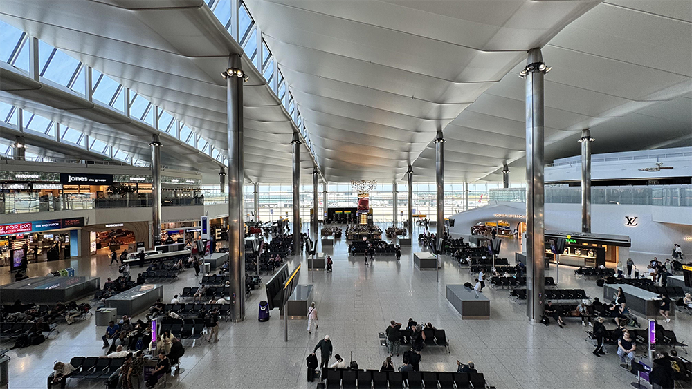 Large, modern airport terminal with high ceilings, glass walls, shops, and seating areas. Many people are waiting, some walking, under bright natural light from large windows.