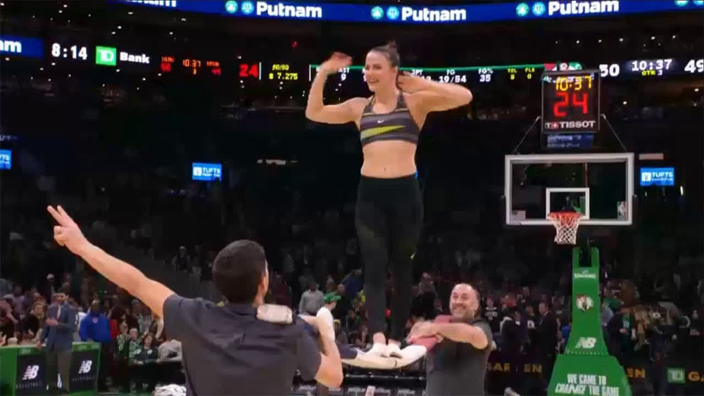 A woman in athletic wear balances on two men’s hands during a basketball game halftime show, with a cheering crowd and scoreboard visible in the background.