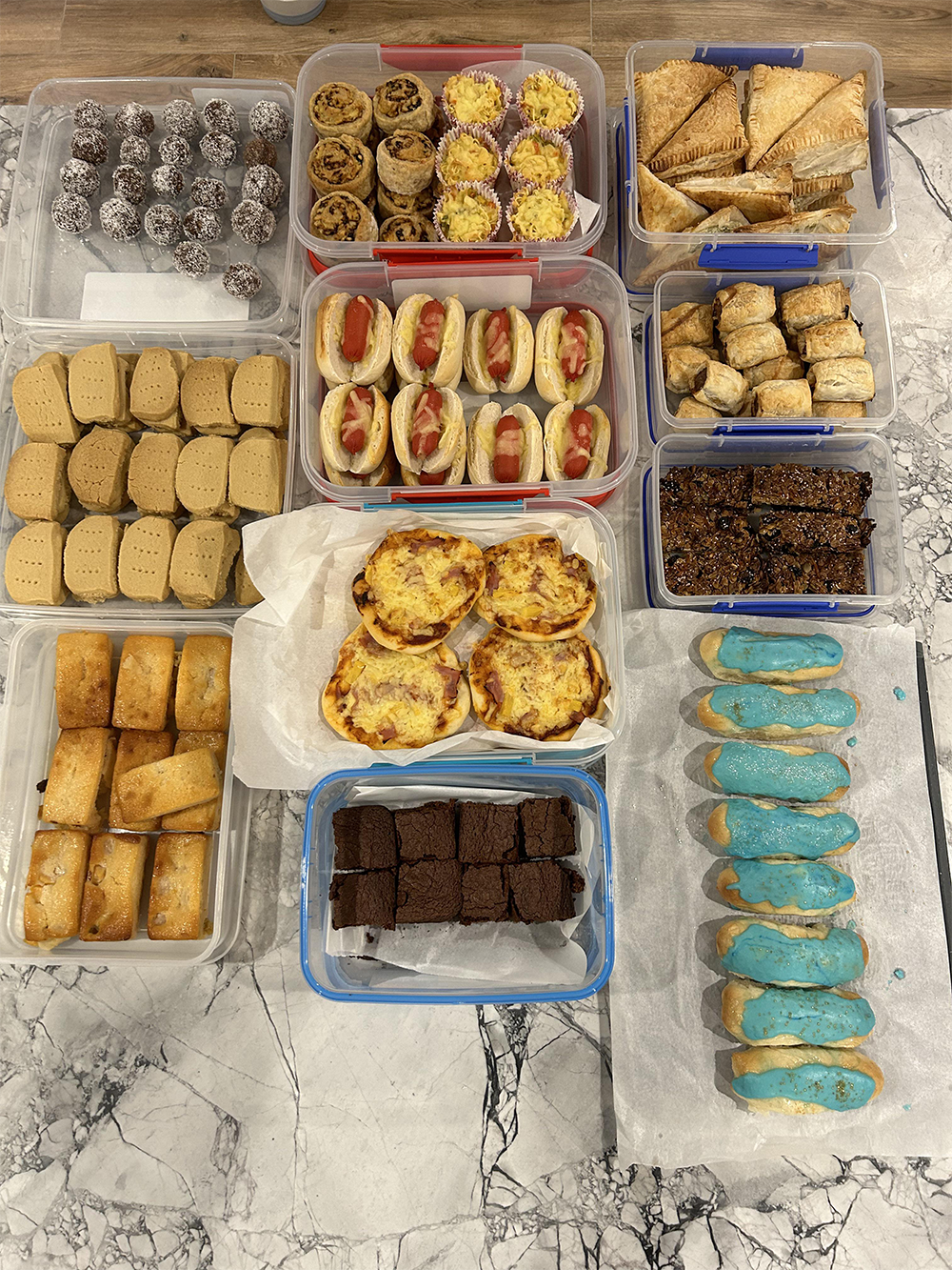 A variety of baked goods, including cookies, bars, pastries, mini pizzas, savory tarts, and blue-iced éclairs, are arranged in containers on a marble countertop.