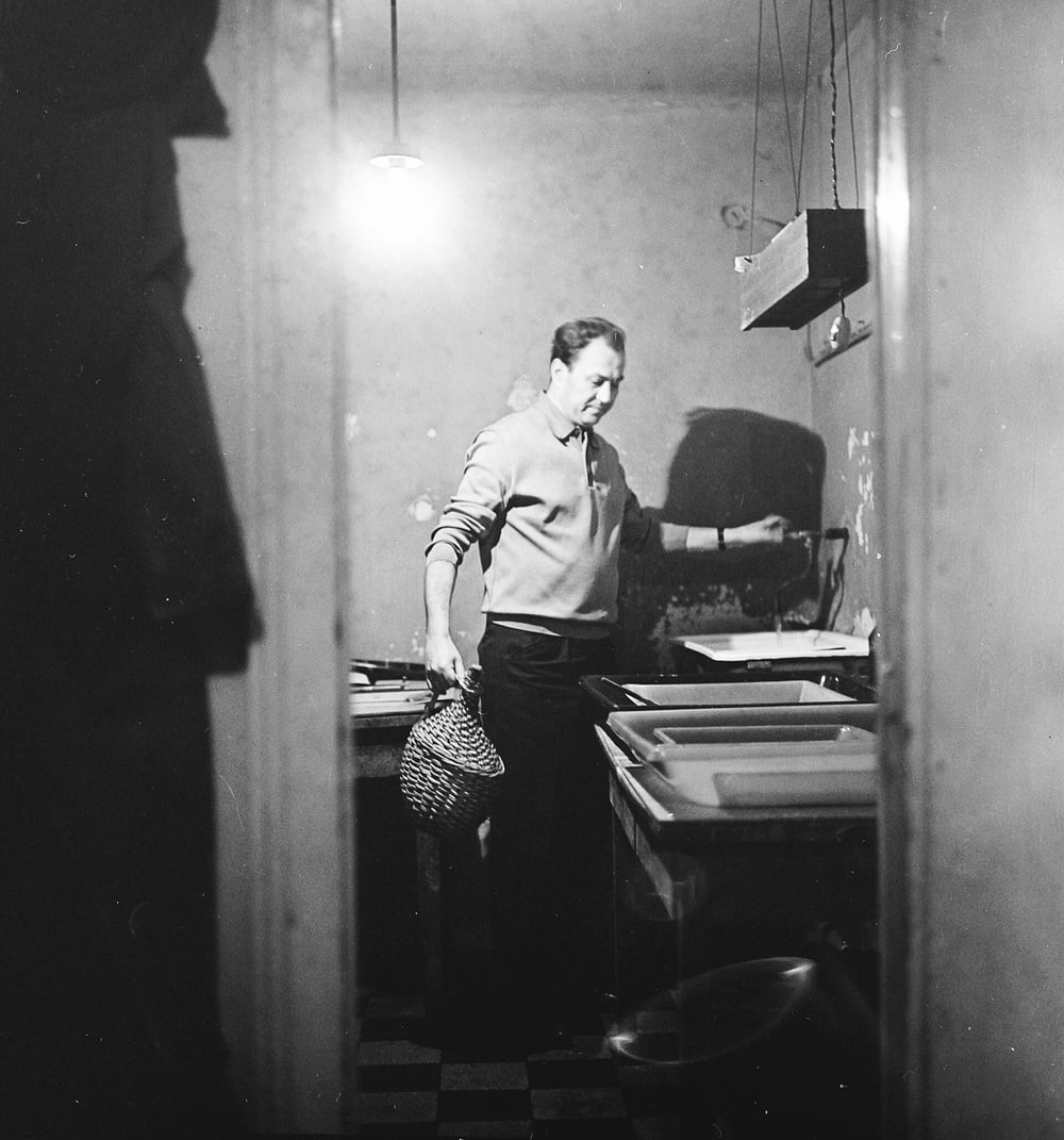 Black and white photo of a person standing in a dimly lit room, holding a basket and looking at trays on a table under a hanging light bulb.